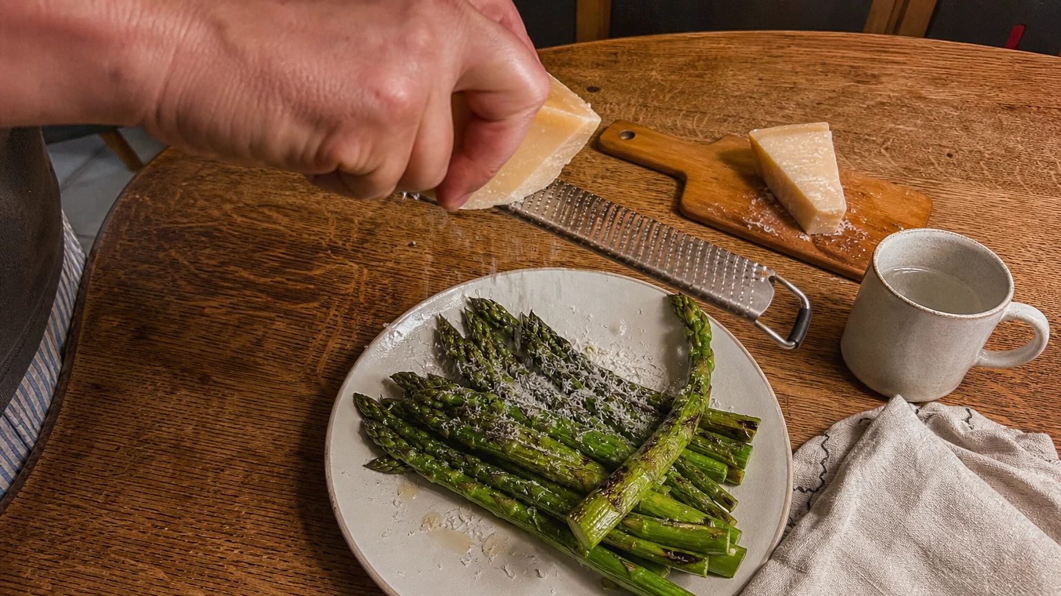 A close-up shot of a hand grating fresh Parmesan cheese over a plate of roasted green asparagus on a rustic wooden dining table.