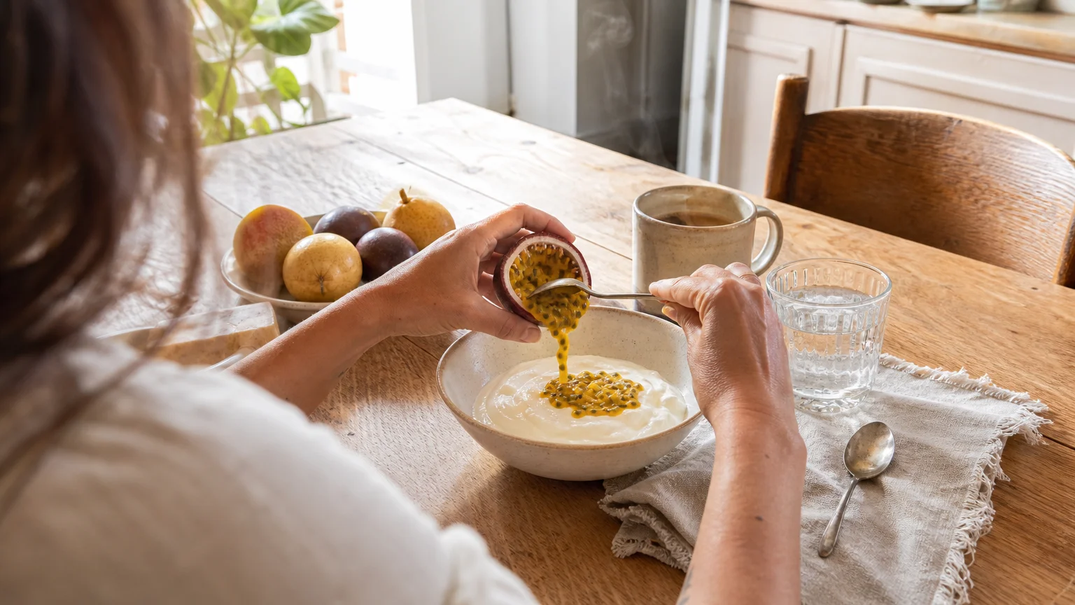 An over-the-shoulder view of a person scooping fresh passion fruit pulp onto a bowl of yogurt at a sunlit wooden kitchen table.