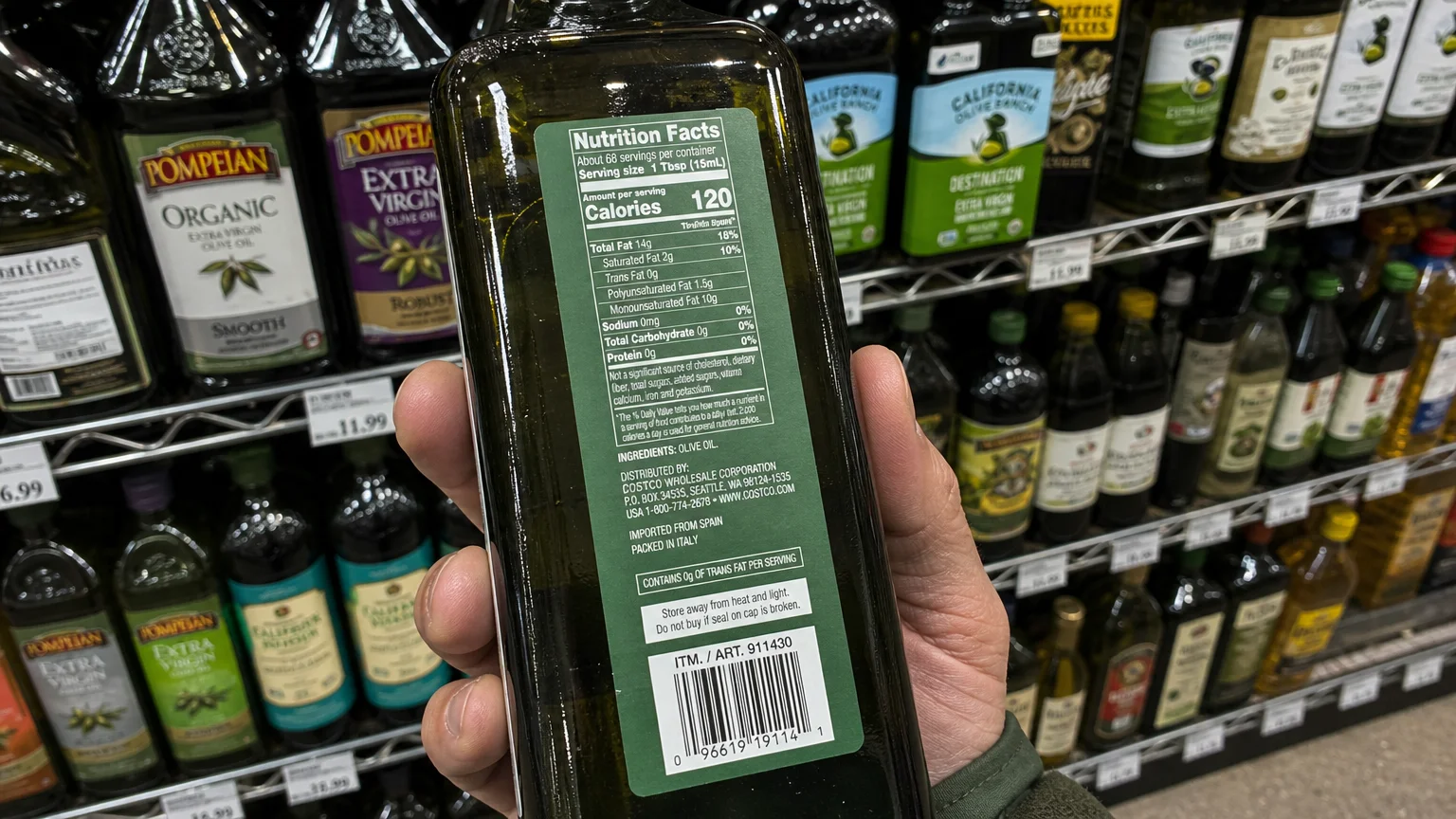First-person view of a hand holding a dark glass olive oil bottle on a grocery store aisle shelf to inspect the back label.