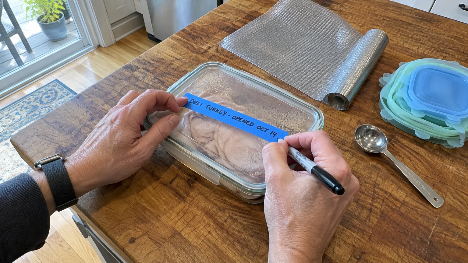 Hands writing an opening date on blue painter's tape atop a glass food storage container filled with sliced deli turkey.