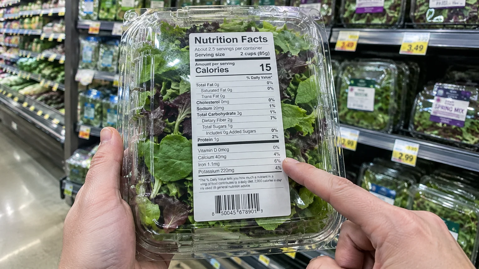 A person holds a plastic container of mixed greens, pointing to the Nutrition Facts label in a grocery aisle.