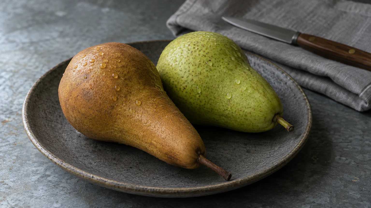 A brown Bosc pear and a green pear with water droplets sit on a dark ceramic plate next to a knife and napkin.