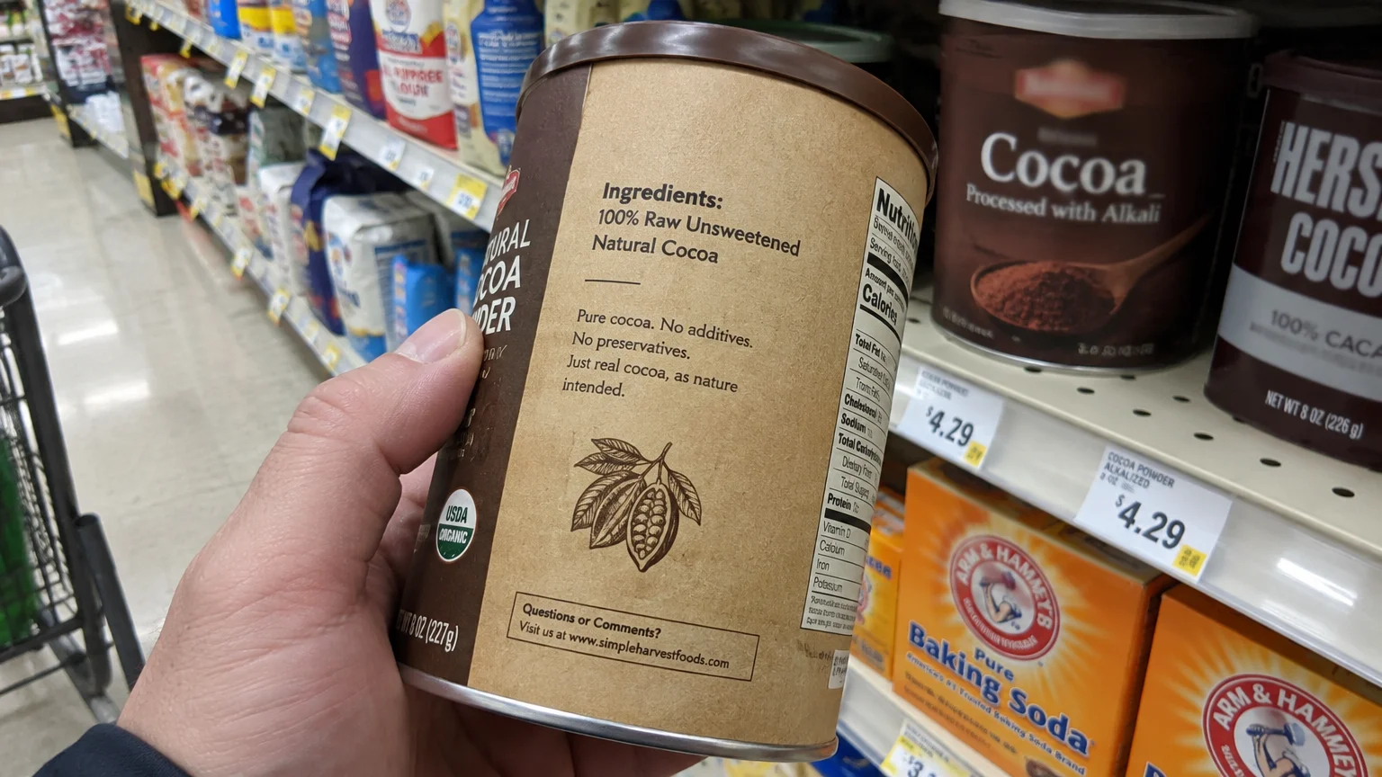Hand holding a natural cocoa canister showing ingredients, with Dutch-processed cocoa powder on a grocery store shelf behind it.